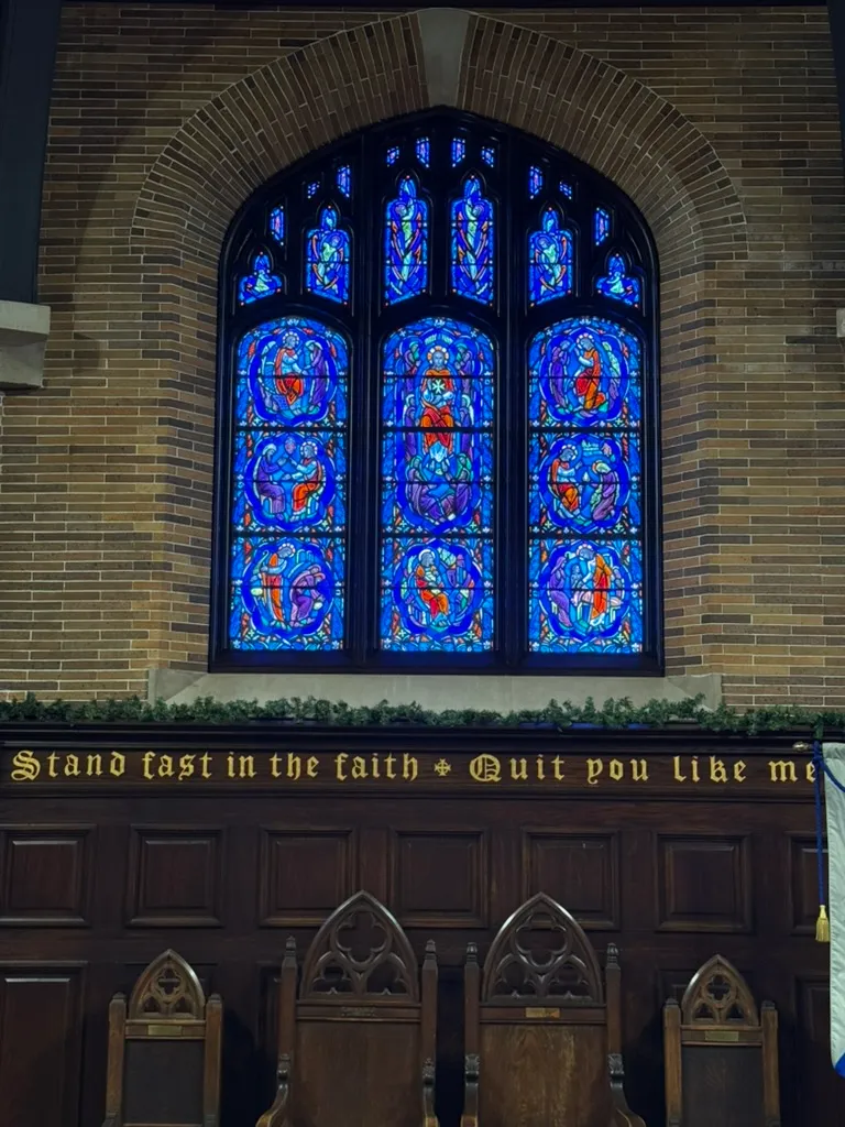 A three-panel cobalt-blue stained-glass window set in a brick wall, above dark wood paneling lettered in gold and carved wooden chairs.