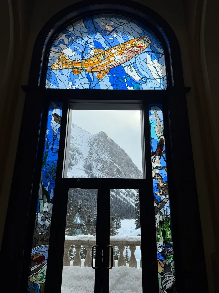 An arched stained-glass window with a golden trout swimming through blue water, framing a snowy mountain through a clear pane below.